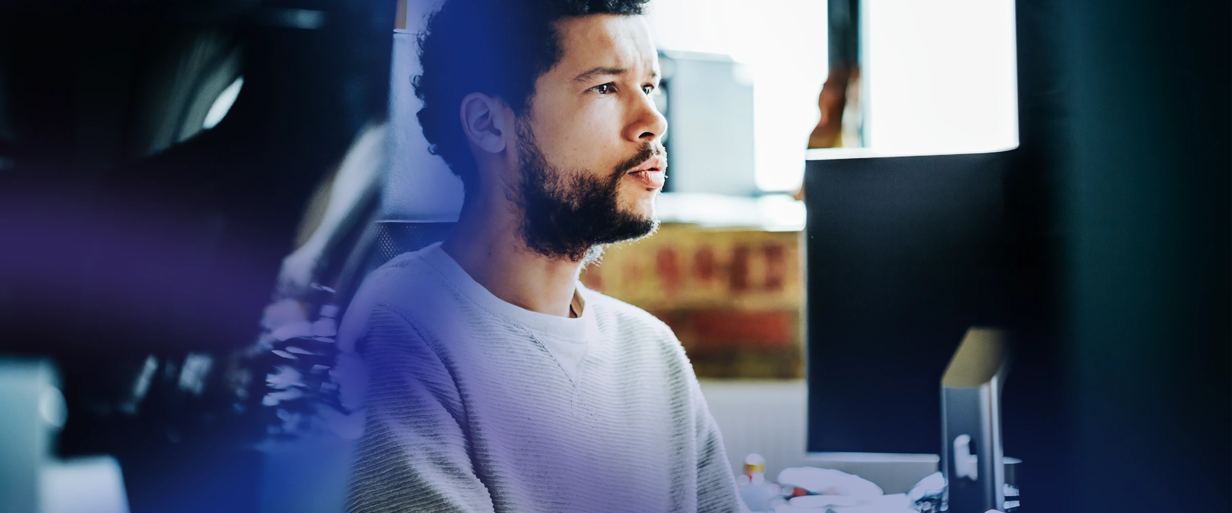 Young man with curly hair and beard wearing a light-colored sweater, sitting and looking attentively at a computer screen in a softly lit office space.