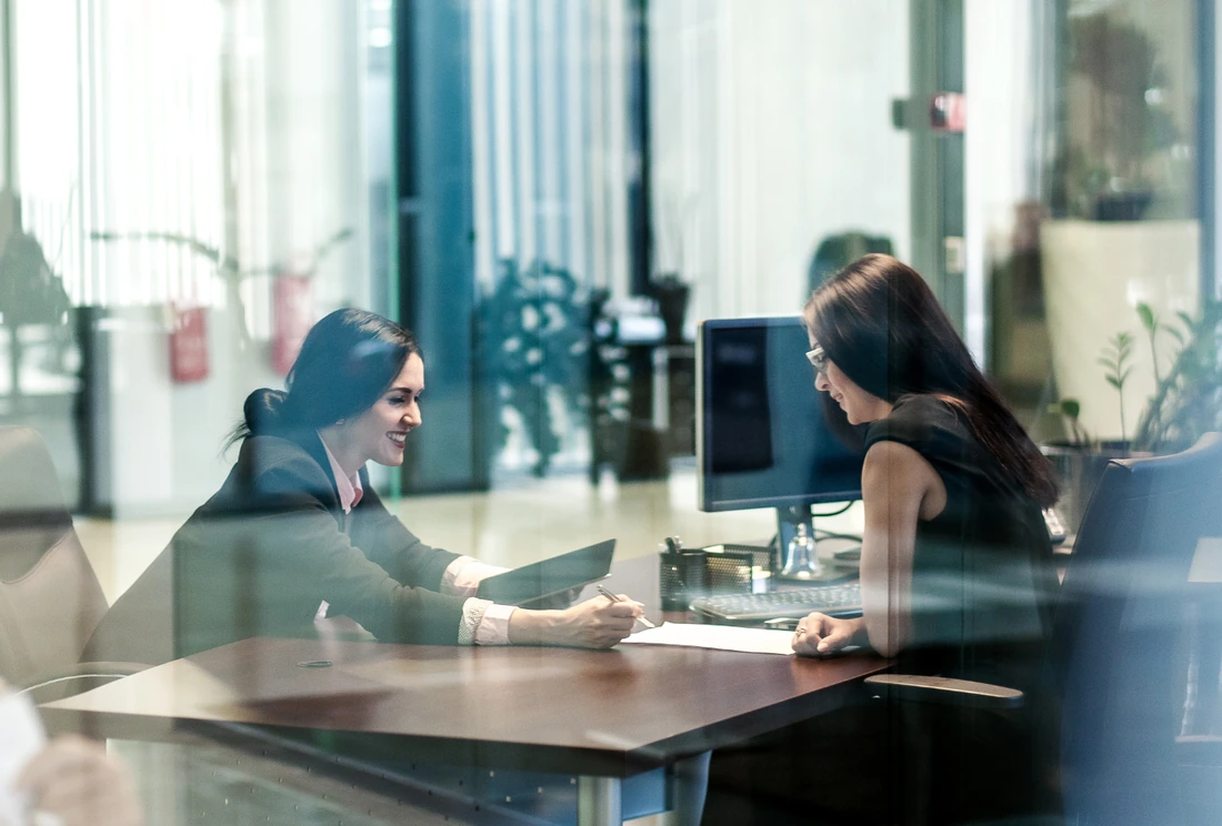 Two women seated at a desk in a modern office, engaged in discussion with one smiling and writing on a document while the other listens attentively; a computer and office supplies are on the desk.
