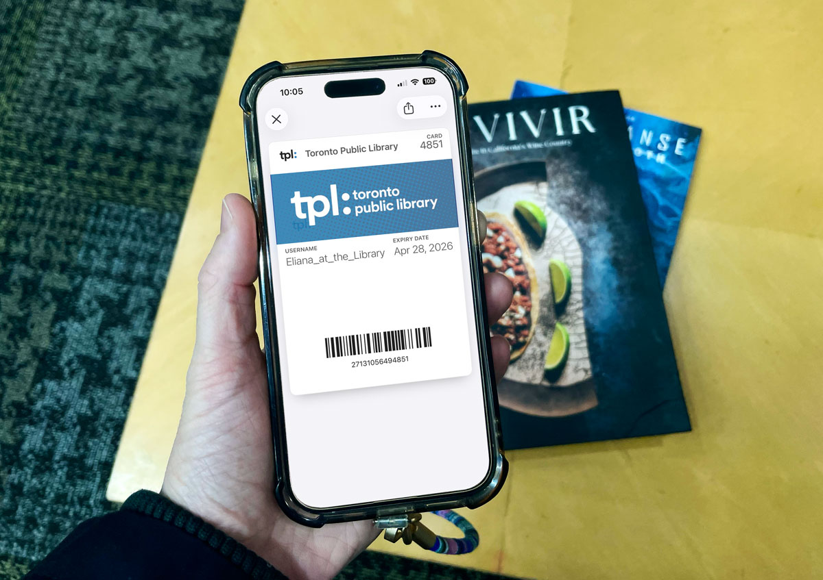 Hand holding a smartphone displaying a Toronto Public Library digital card with barcode and expiration date, resting above books on a table inside the library.