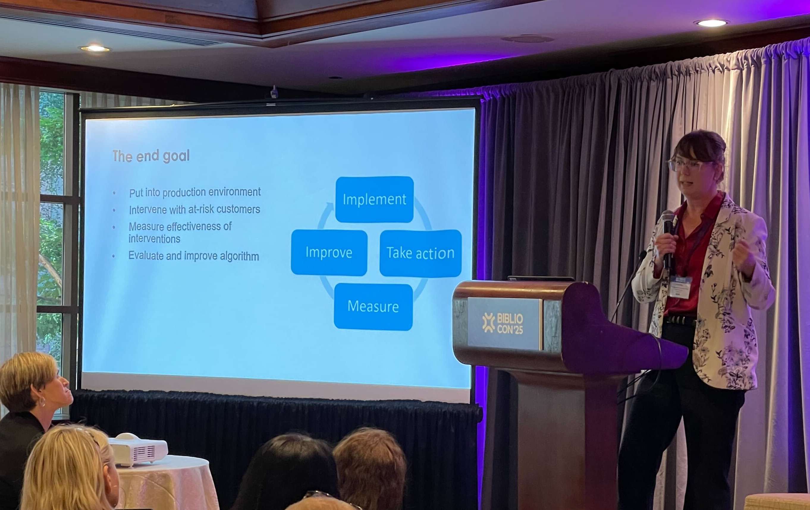 Presenter speaking at a podium during a BiblioCon session, with a slide titled “The end goal” showing steps to implement, measure, improve, and take action while attendees watch from the audience.