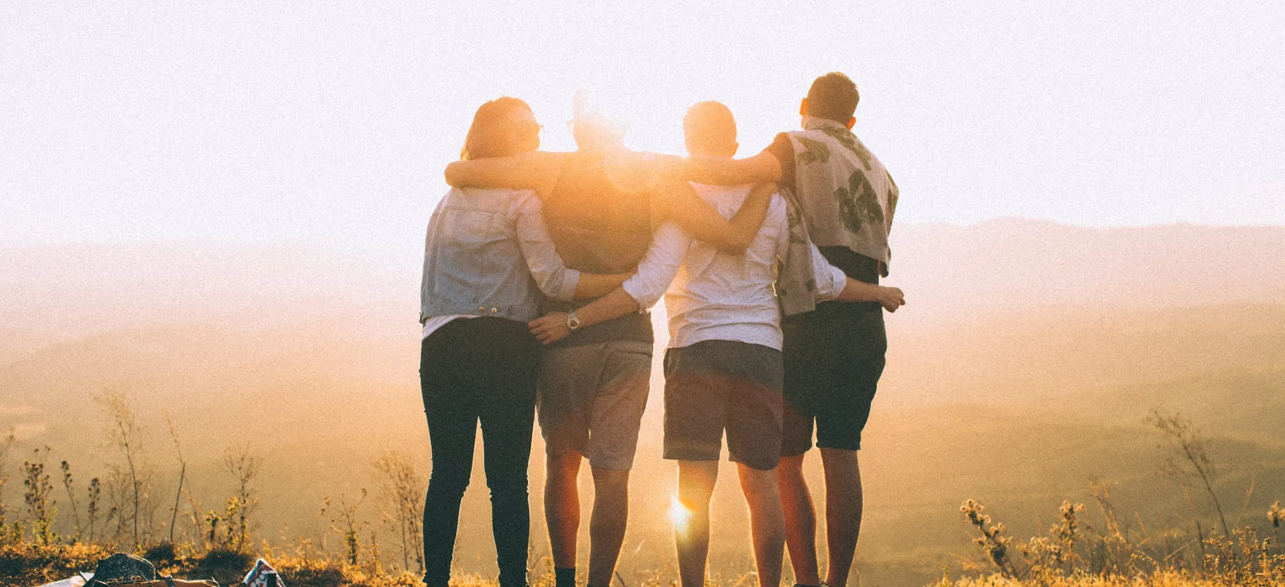 A group of friends with their arms around each other's shoulders looking into the sunset.