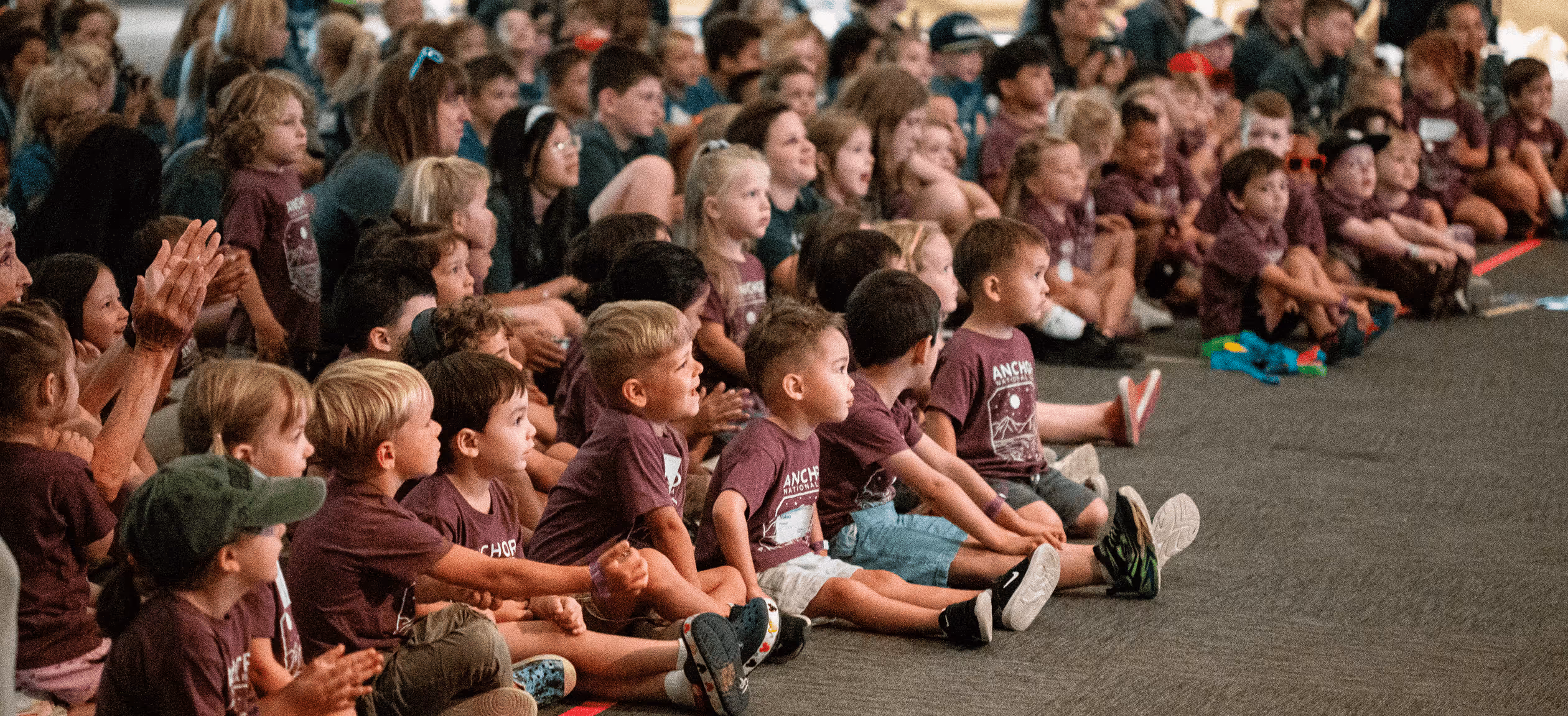 A large group of children sitting on the floor, attentively watching something off-camera, many wearing matching maroon shirts.