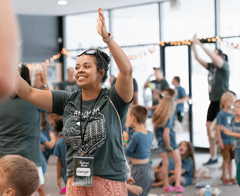Woman with a name tag raising her hands and smiling amid a group of children and adults in an indoor activity space.