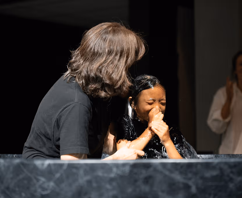 Person in black shirt assisting another person who is getting baptized and covering their face from water.