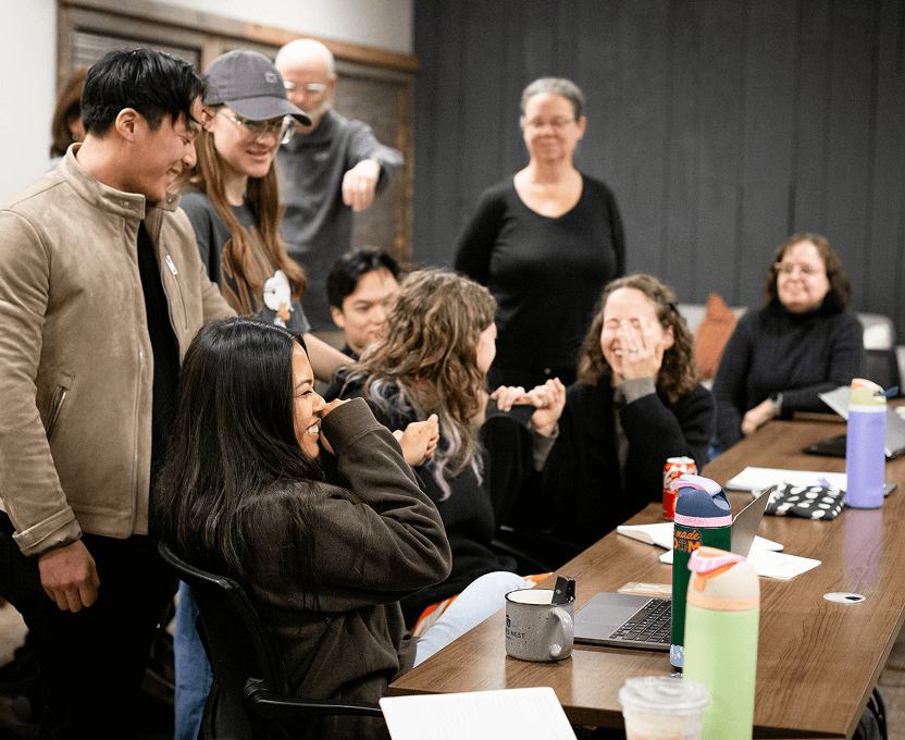 A group of diverse people gathered around a conference table, laughing and engaging in a lively discussion with laptops and drinks on the table.