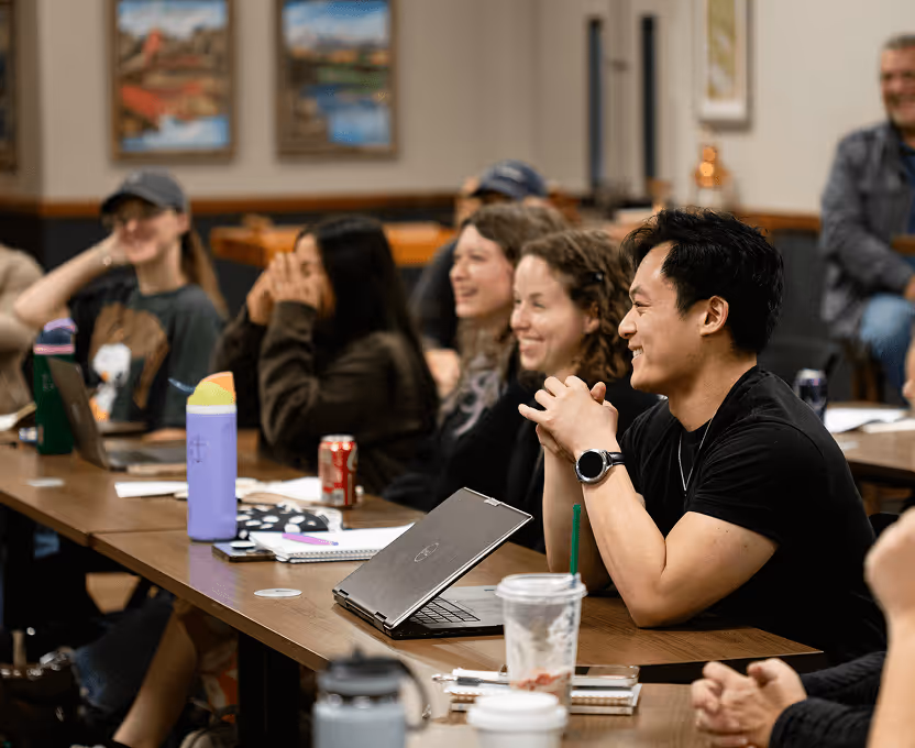 Group of people smiling and listening attentively in a classroom or meeting room setting with laptops and notebooks on the table.
