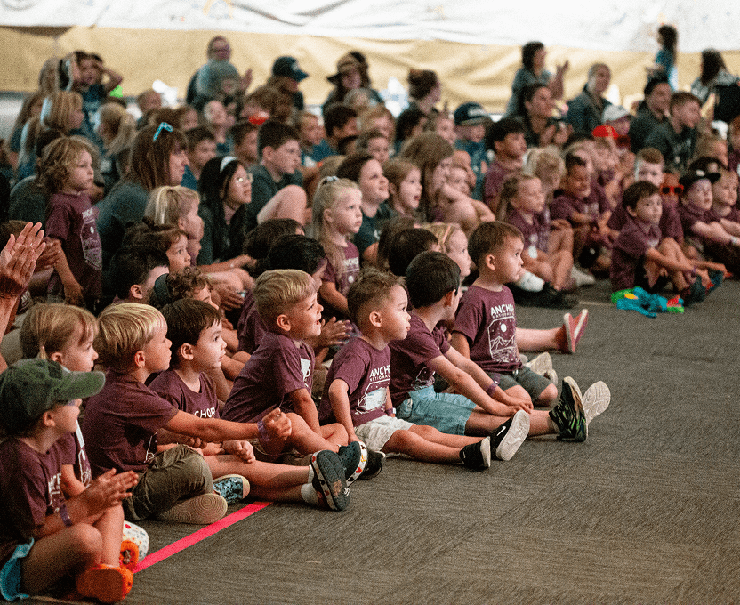 Large group of children seated on the floor indoors, attentively watching something to the right, many wearing matching maroon t-shirts.