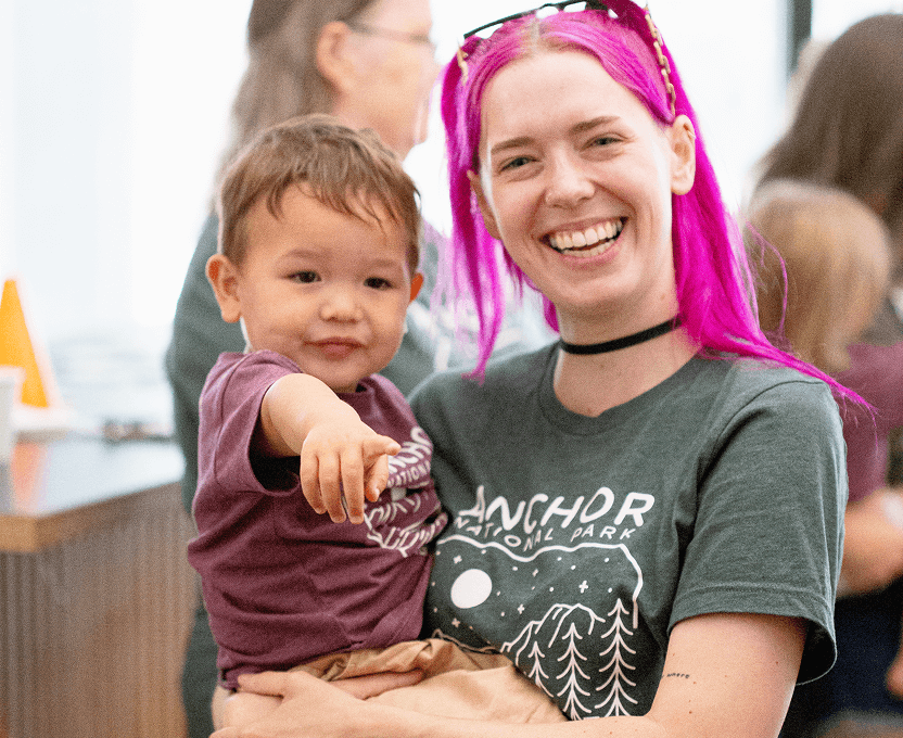 Smiling woman with bright pink hair holding a young child who is pointing forward, both wearing matching dark t-shirts.