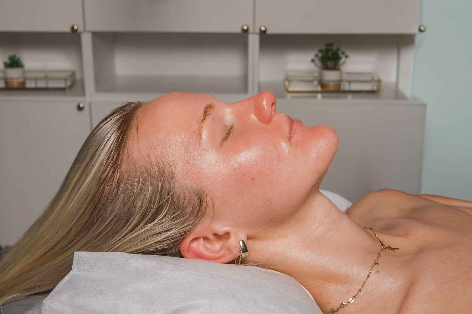 A woman with glowing, freshly treated skin lies on a spa treatment bed with her eyes closed, appearing relaxed.