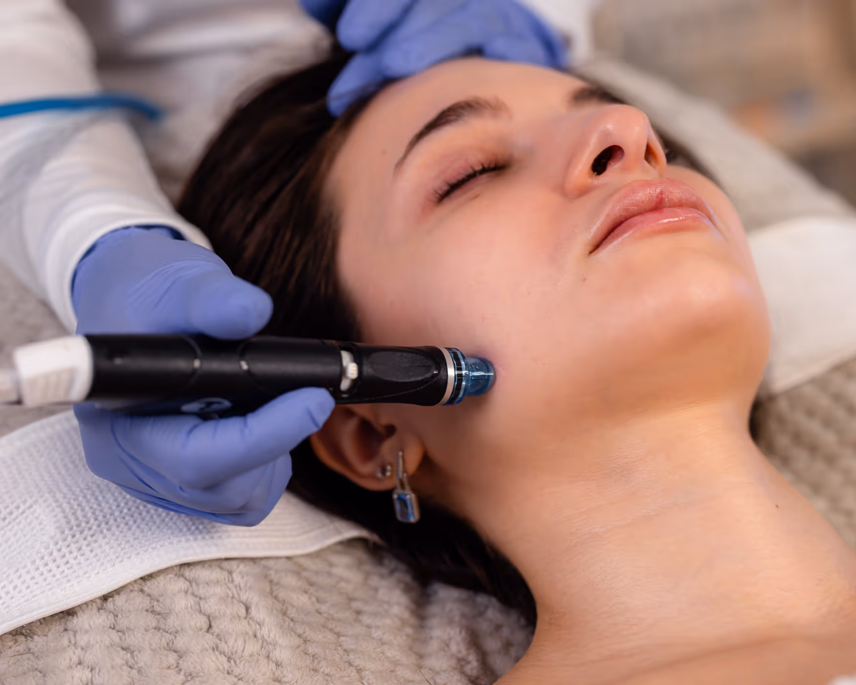 A licensed esthetician wearing blue gloves performs a professional facial treatment using a HydraFacial device on a relaxed female client lying on a treatment bed.