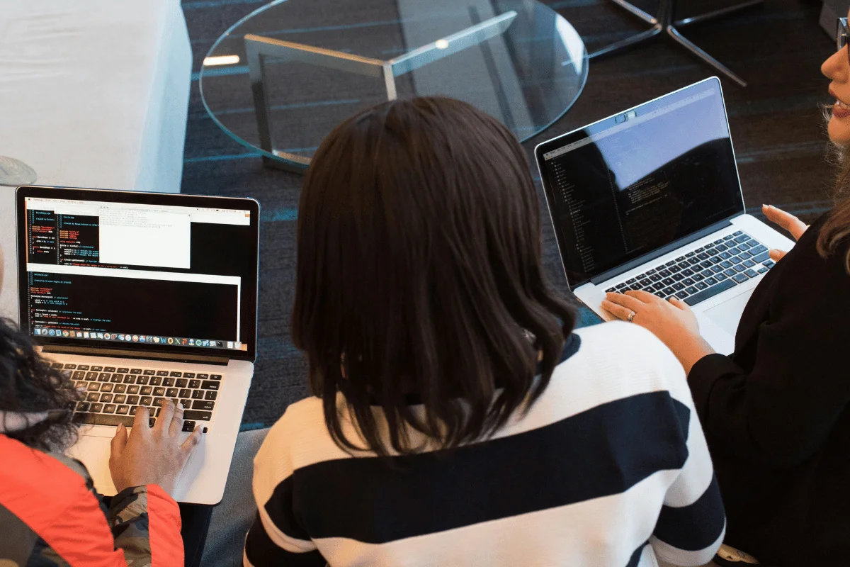 A group of three people sitting side-by-side, working on laptops with programming code on their screens.