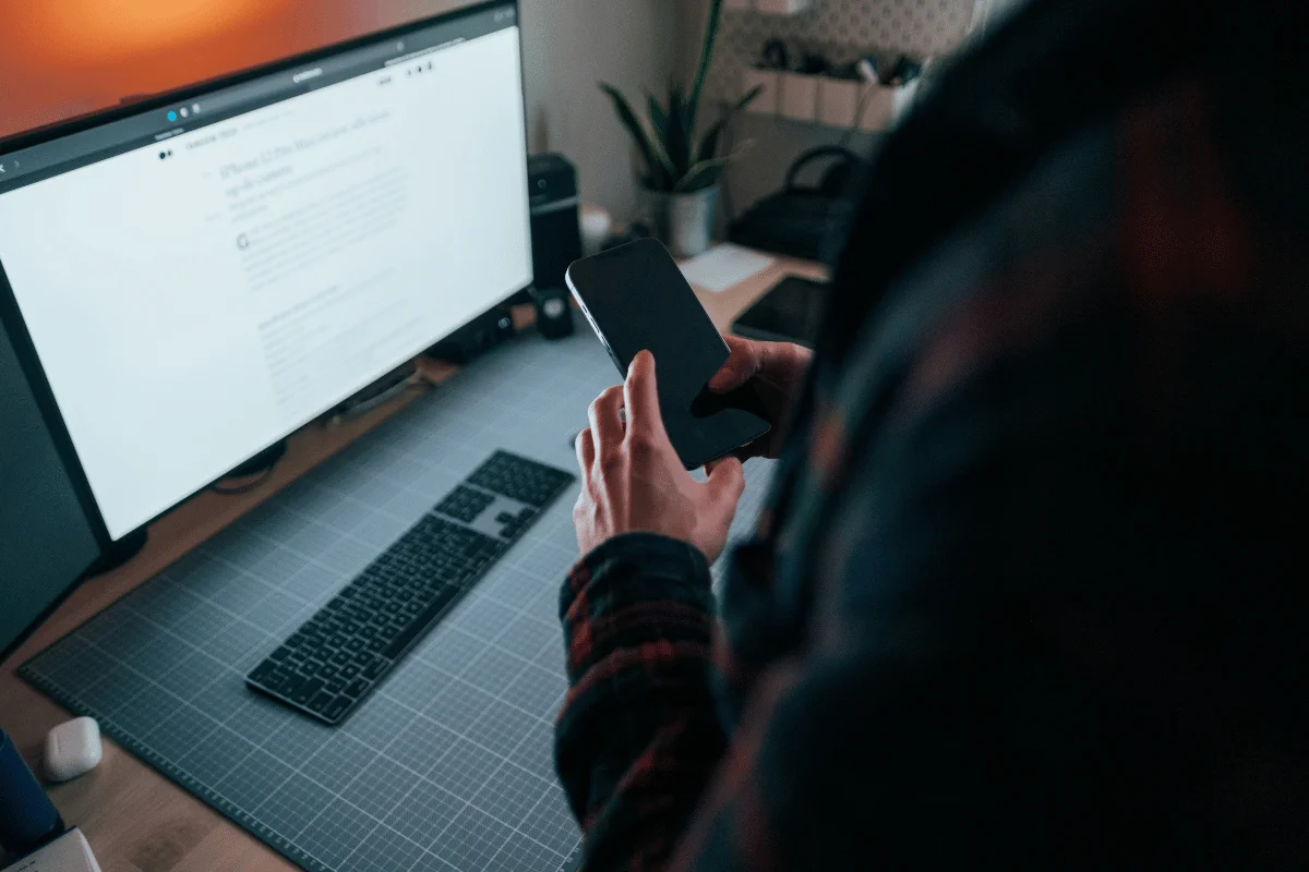 Person holding smartphone while working at desktop computer.
