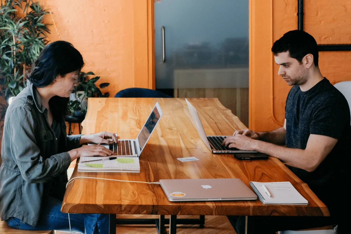 Two coworkers working on laptops at wooden office table.