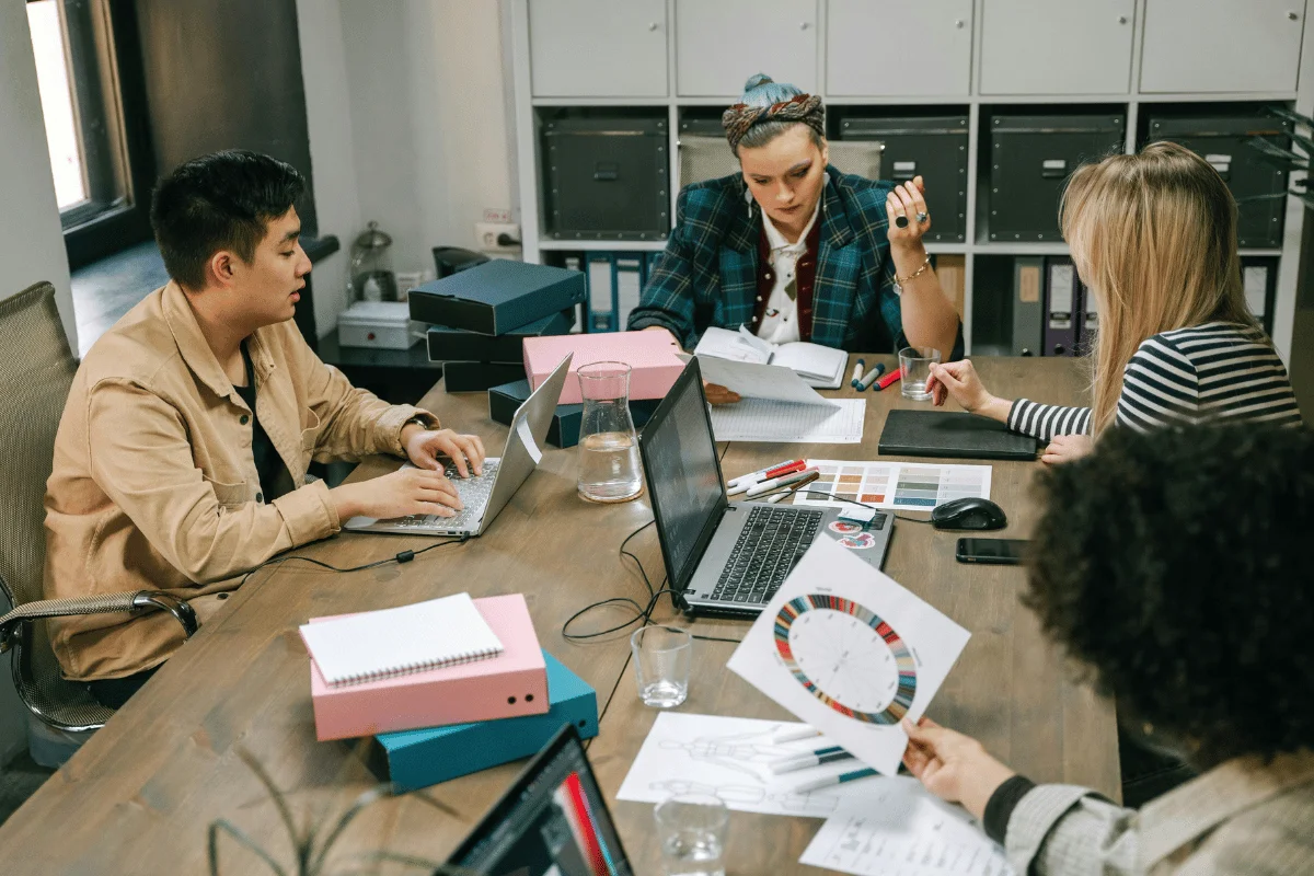 Four people collaborate at a wooden table cluttered with laptops, papers, and color charts. The atmosphere is focused and creative.