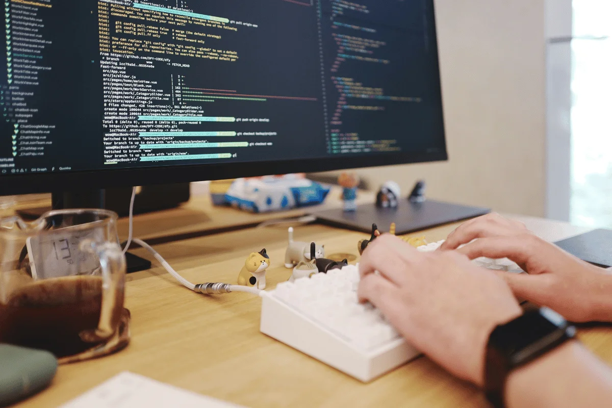 Developer typing code on desktop monitor with coffee and cat figurines on desk.