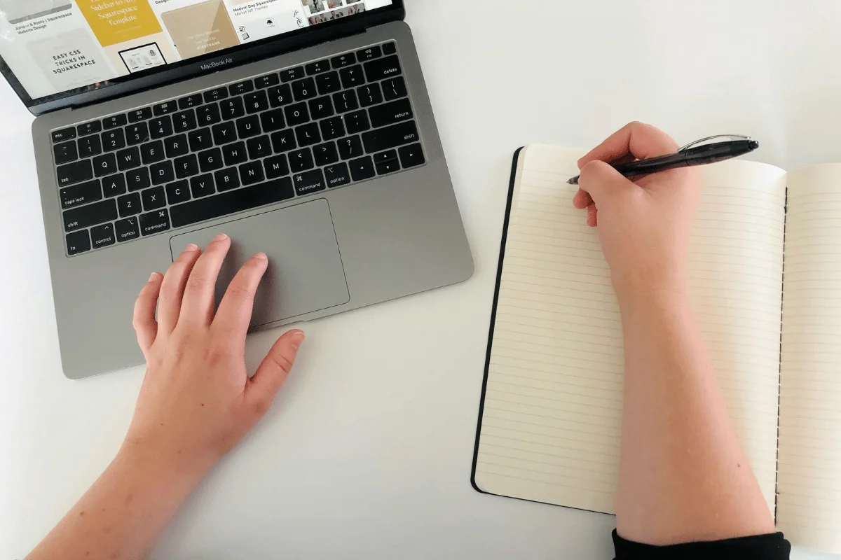 A person's hand using a MacBook Air, while the other hand writes in a lined notebook on a white desk.