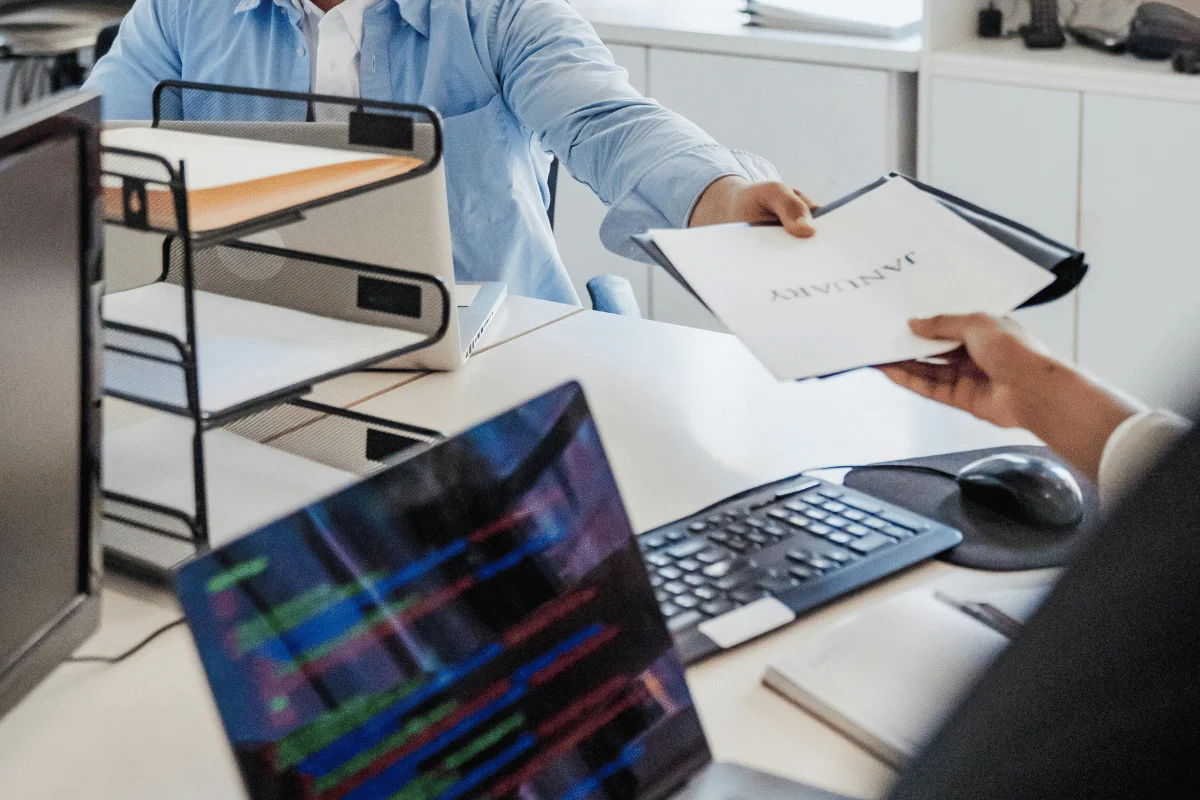 A person in a blue shirt hands a document titled "January" to another person at a desk with laptops and office supplies.