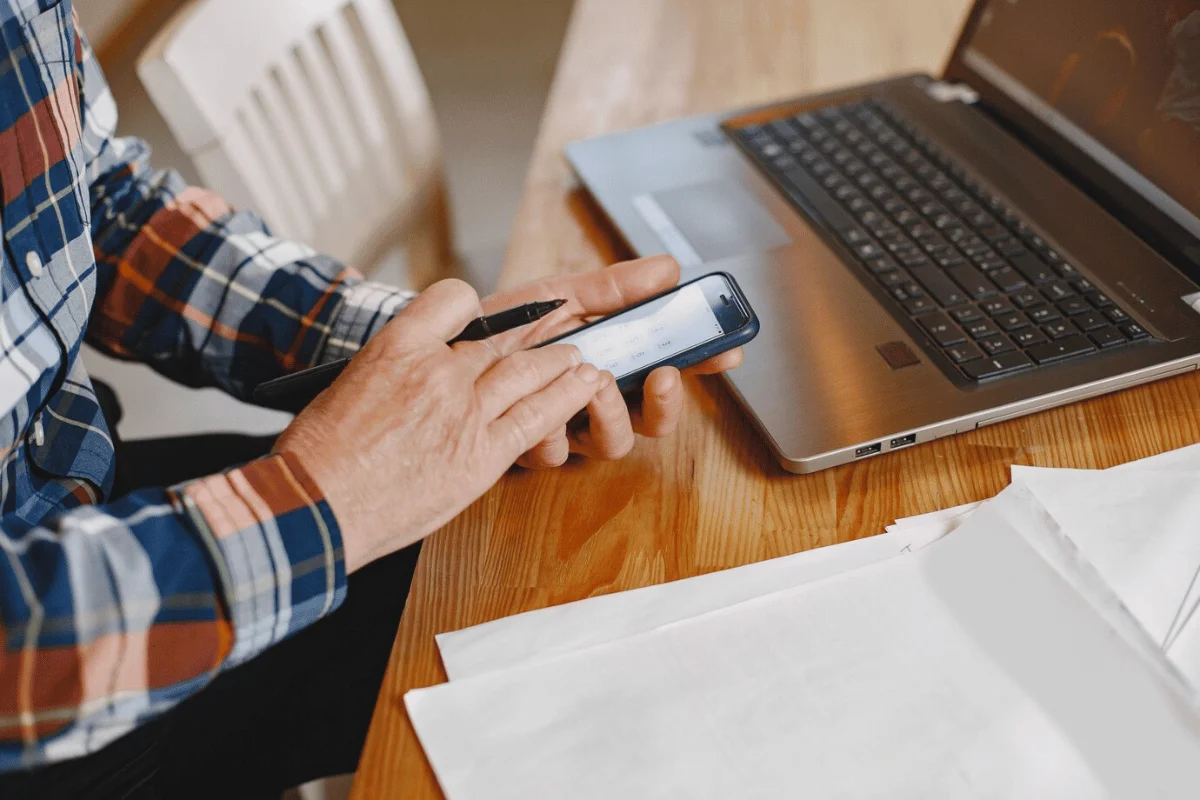 A person using a smartphone while holding a pen next to a laptop on a wooden table.