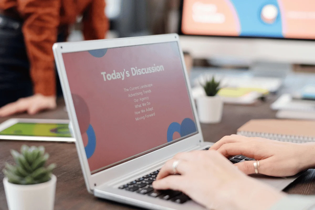 A person types on a laptop displaying a presentation titled "Today's Discussion" in a modern office setting with plants and notes.