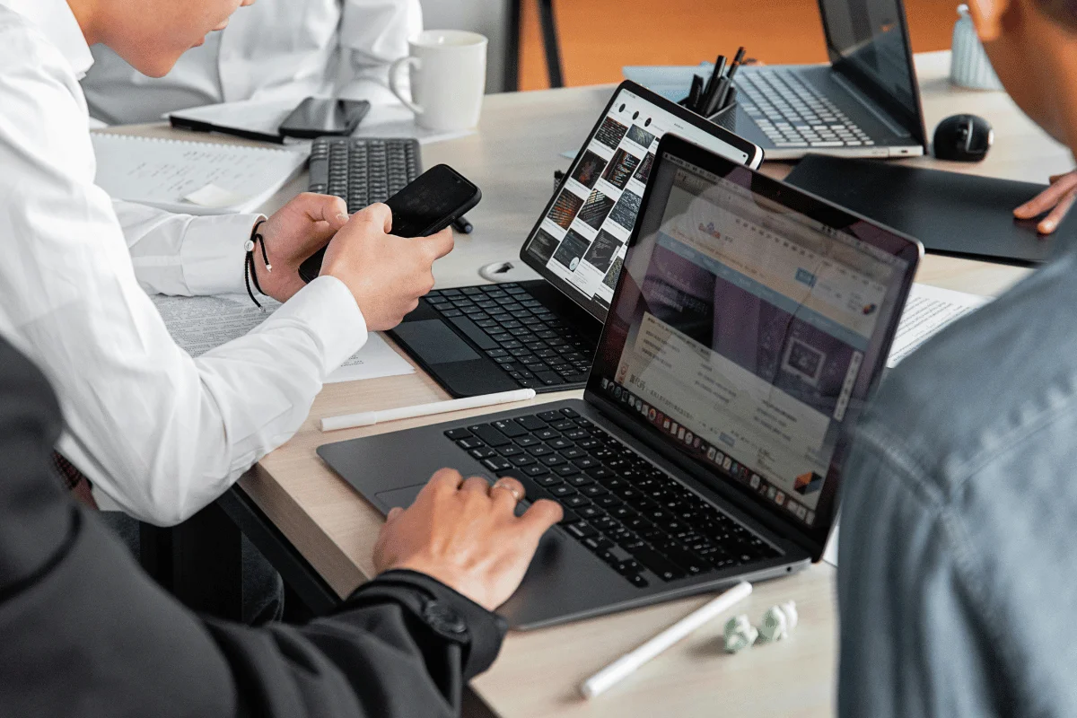 A close-up view of hands using a laptop and smartphone during a meeting, surrounded by papers and office supplies.