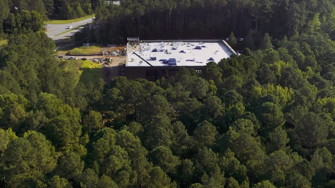 A white-roofed building under construction surrounded by dense green forest and adjacent to a road.