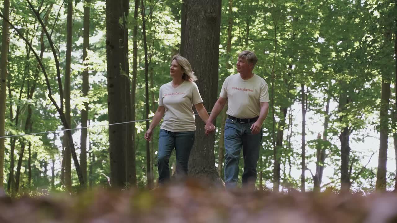 Middle-aged couple holding hands and walking in a sunlit forest with green trees.