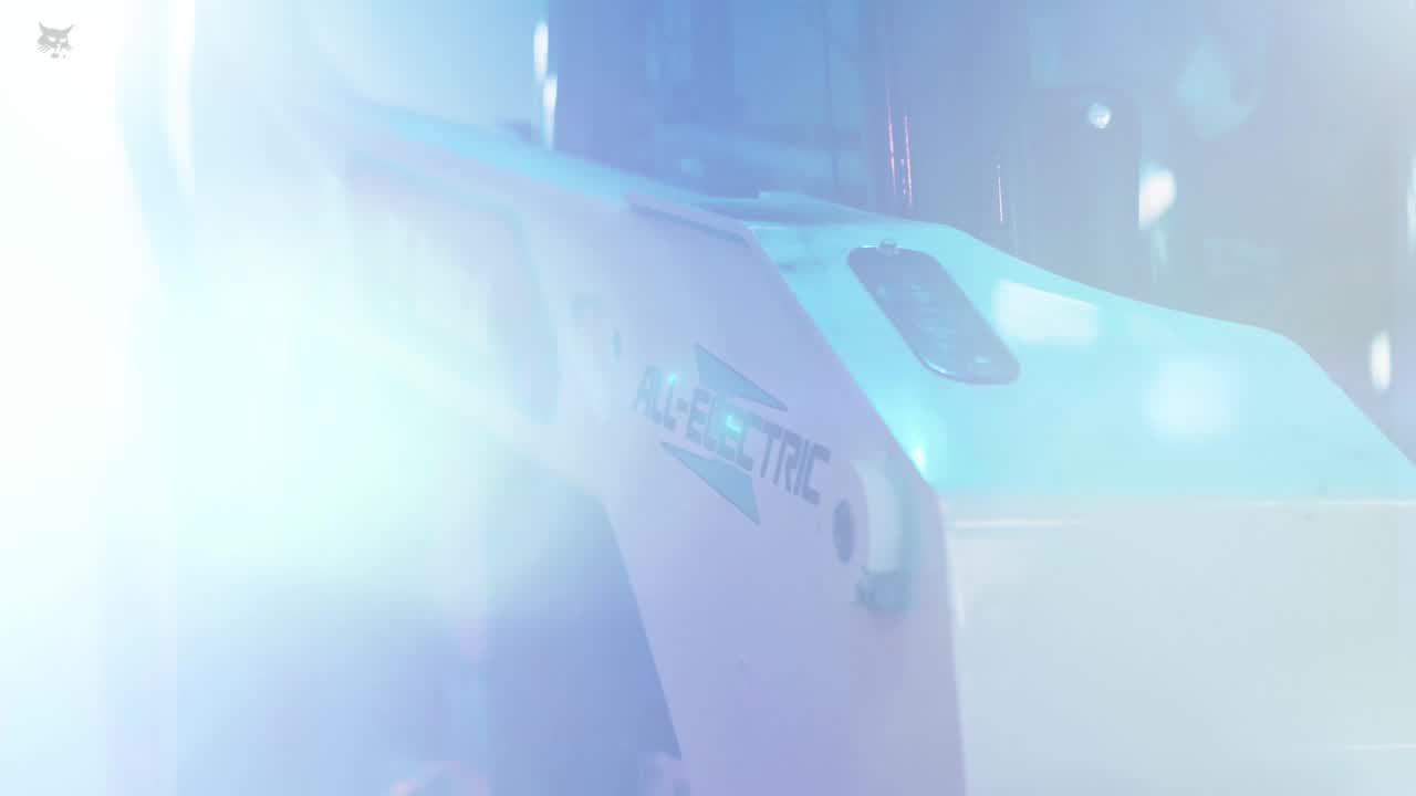 Bobcat all-electric compact track loader scooping dirt inside a dimly lit warehouse with overhead lights.