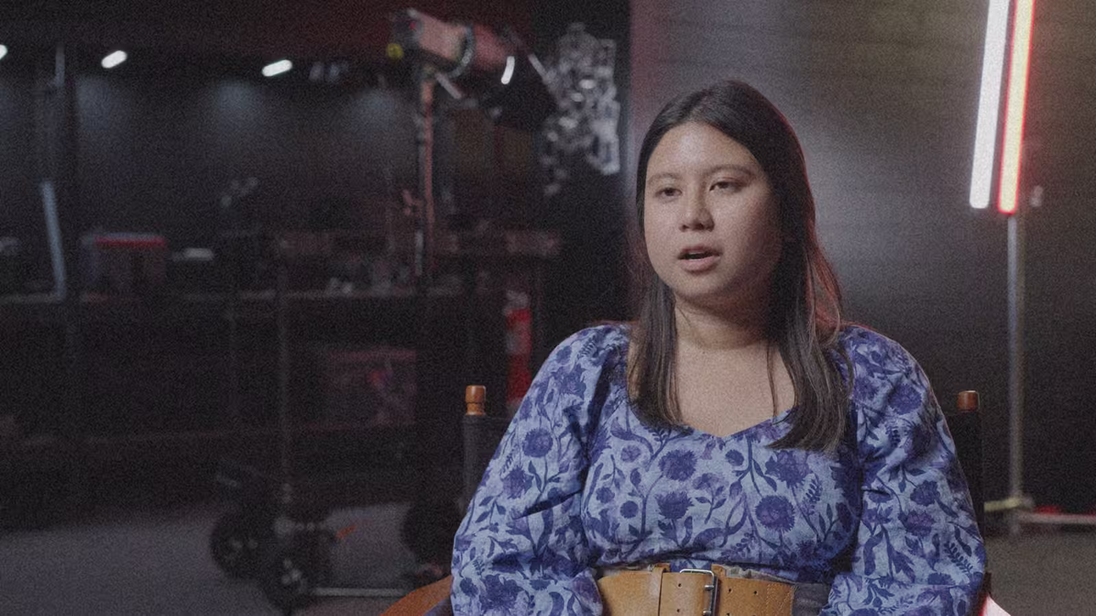 Young woman with long hair, wearing a blue floral blouse and a brown belt, sits in a dimly lit room with filmmaking equipment in the background.