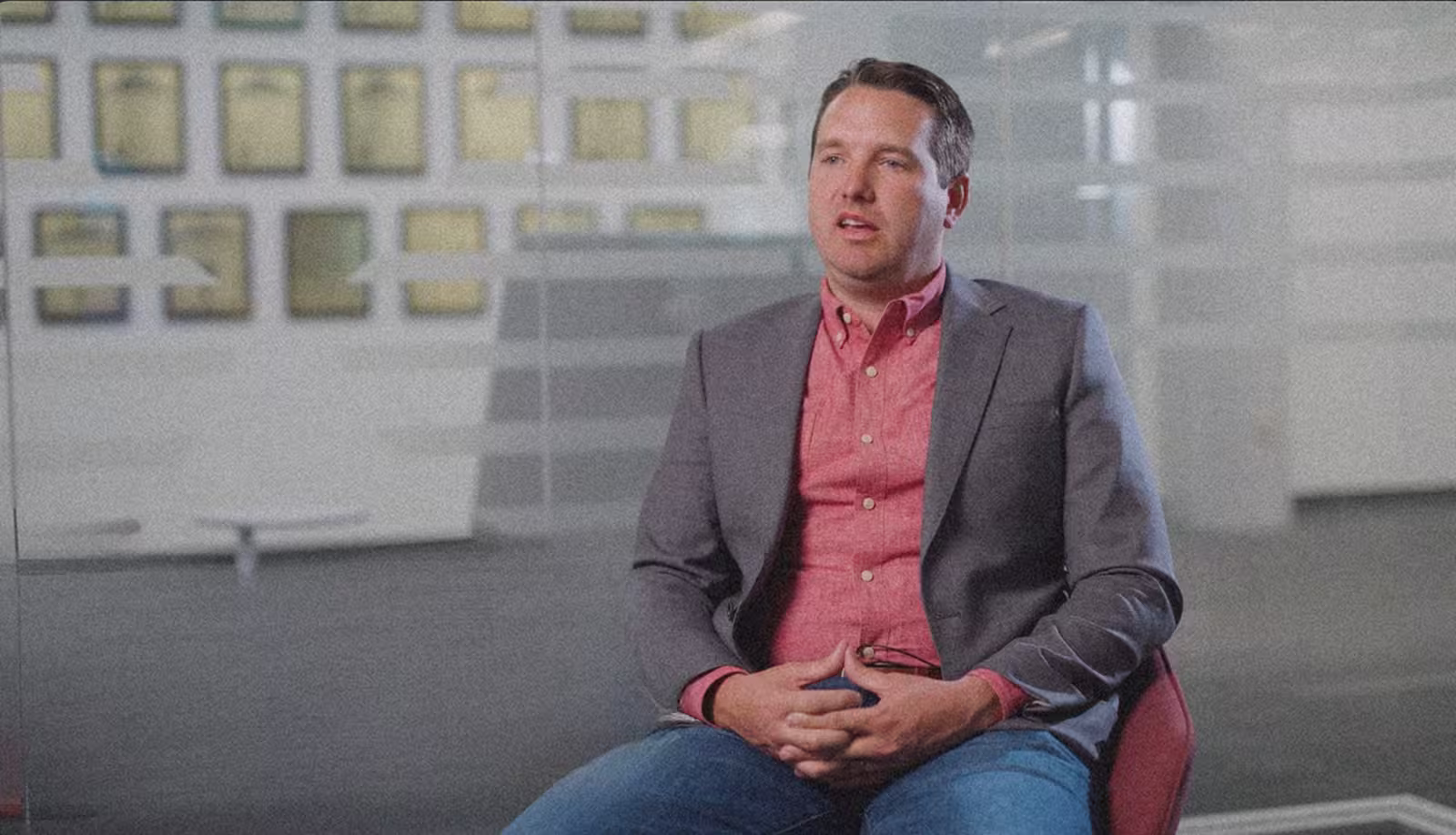 A man in a suit jacket and red shirt sits in an office environment with frosted glass walls and framed pictures. The setting is professional and formal.