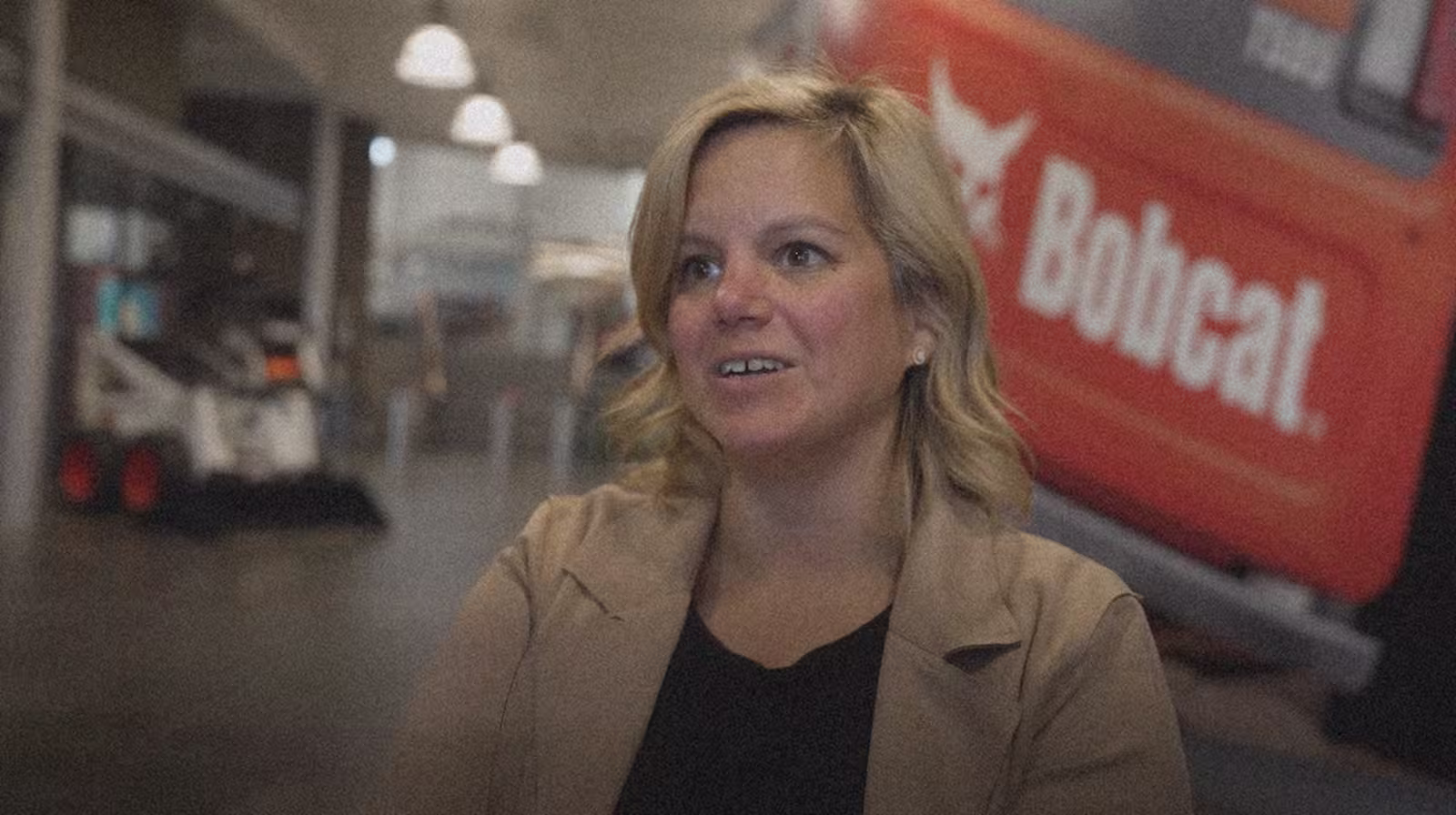 A woman with blonde hair smiles warmly indoors. The background includes industrial equipment and a blurred "Bobcat" logo, creating a professional atmosphere.