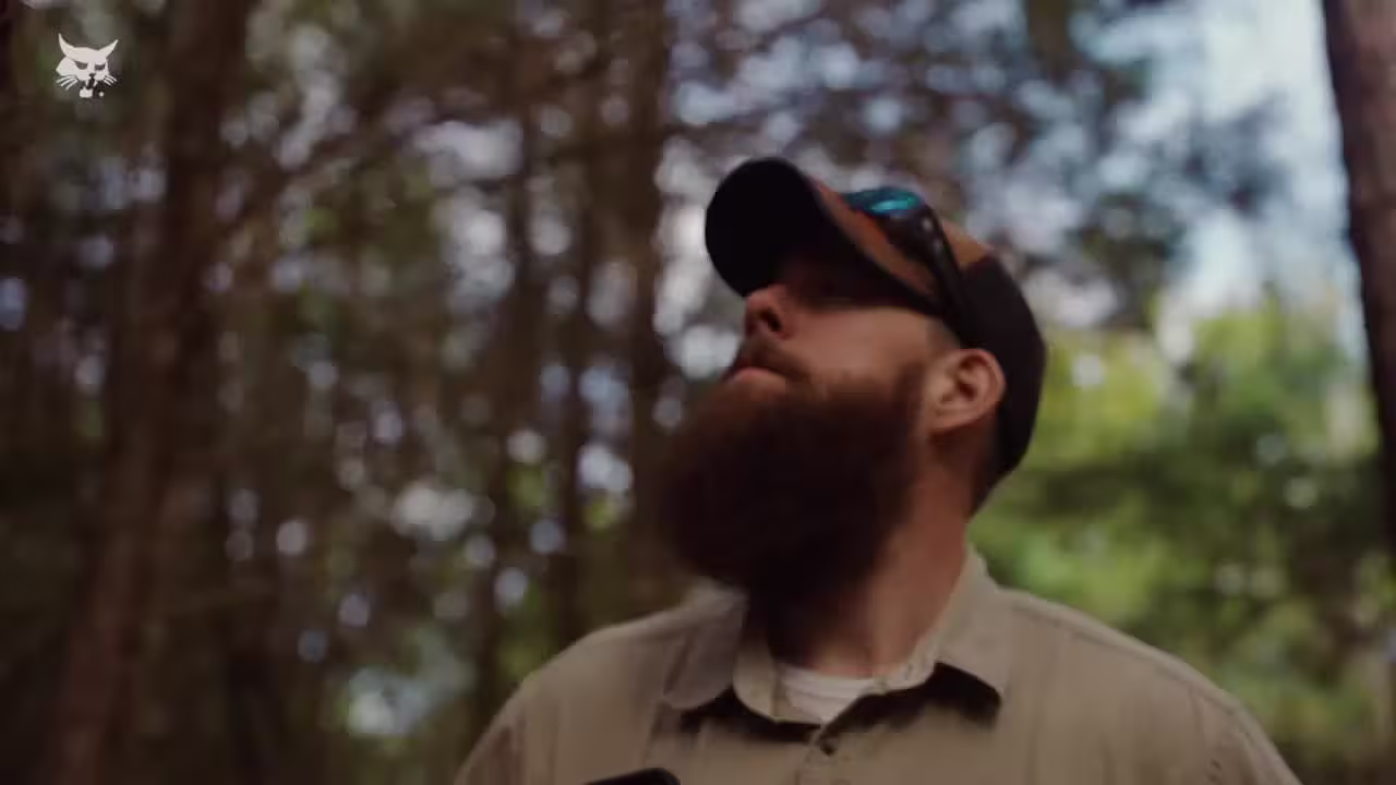 A man with a beard and cap gazes upward in a forest. Dappled sunlight filters through the trees, creating a serene and contemplative atmosphere.