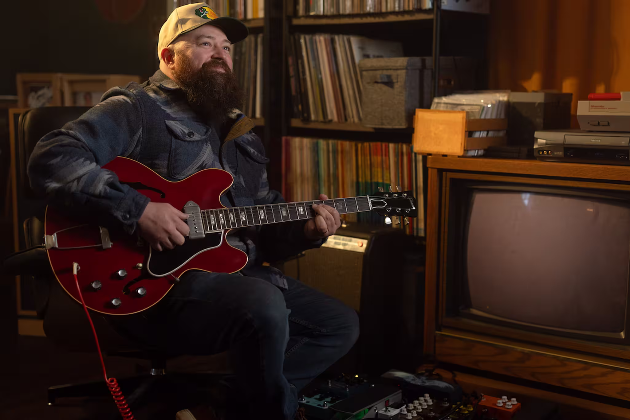 A bearded man wearing a cap smiles while playing a red electric guitar, seated next to a vintage television and shelves of records.