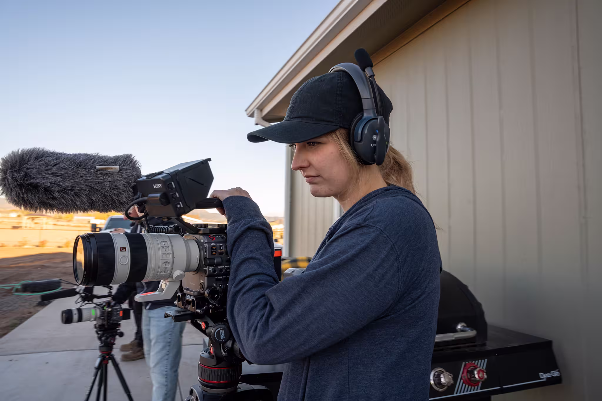 A person in a dark sweatshirt and cap, wearing headphones, operates a professional camera with a large lens and microphone outside a shed.