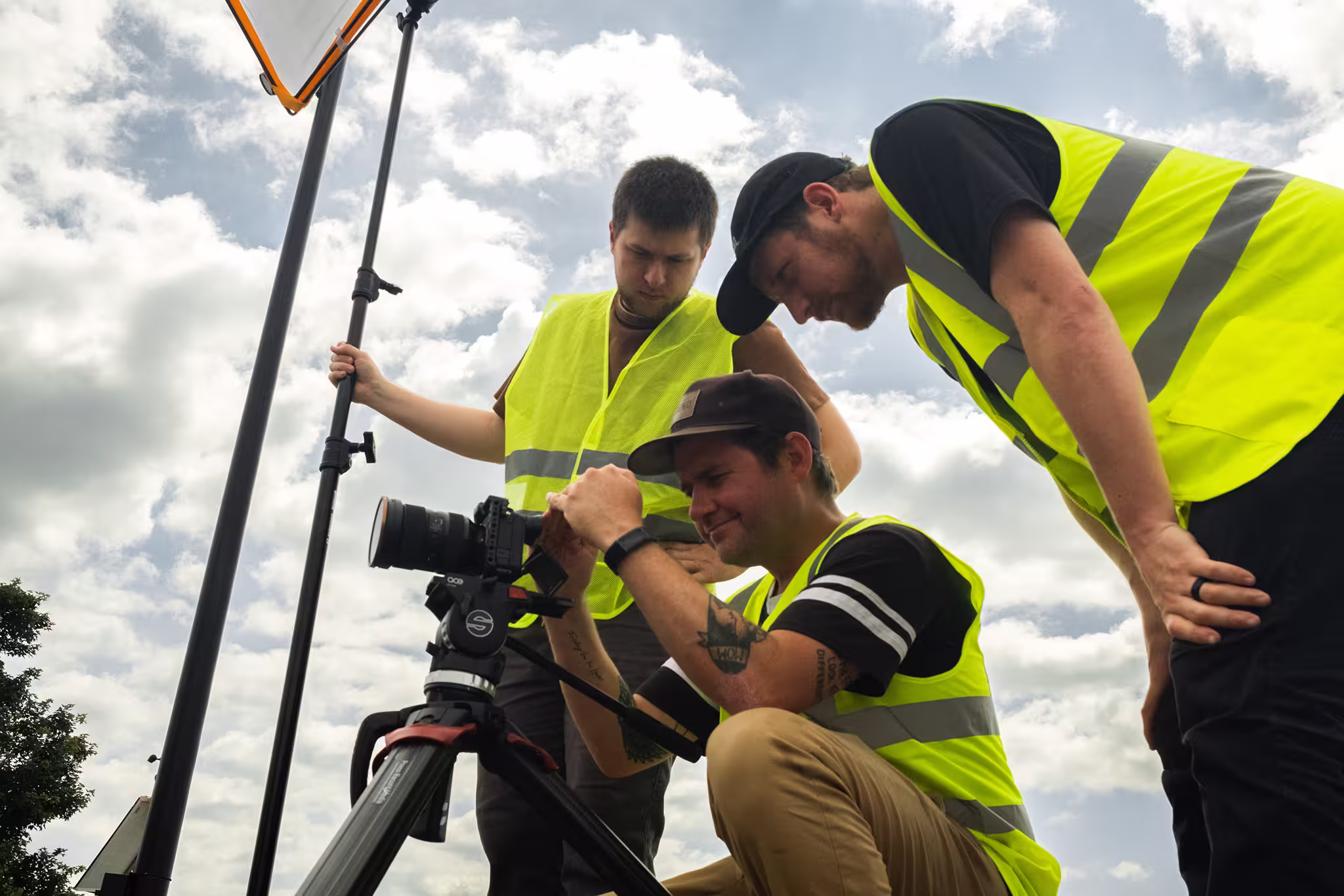 Four men in high-visibility safety vests gather around a camera on a tripod, reviewing footage together under a cloudy sky.