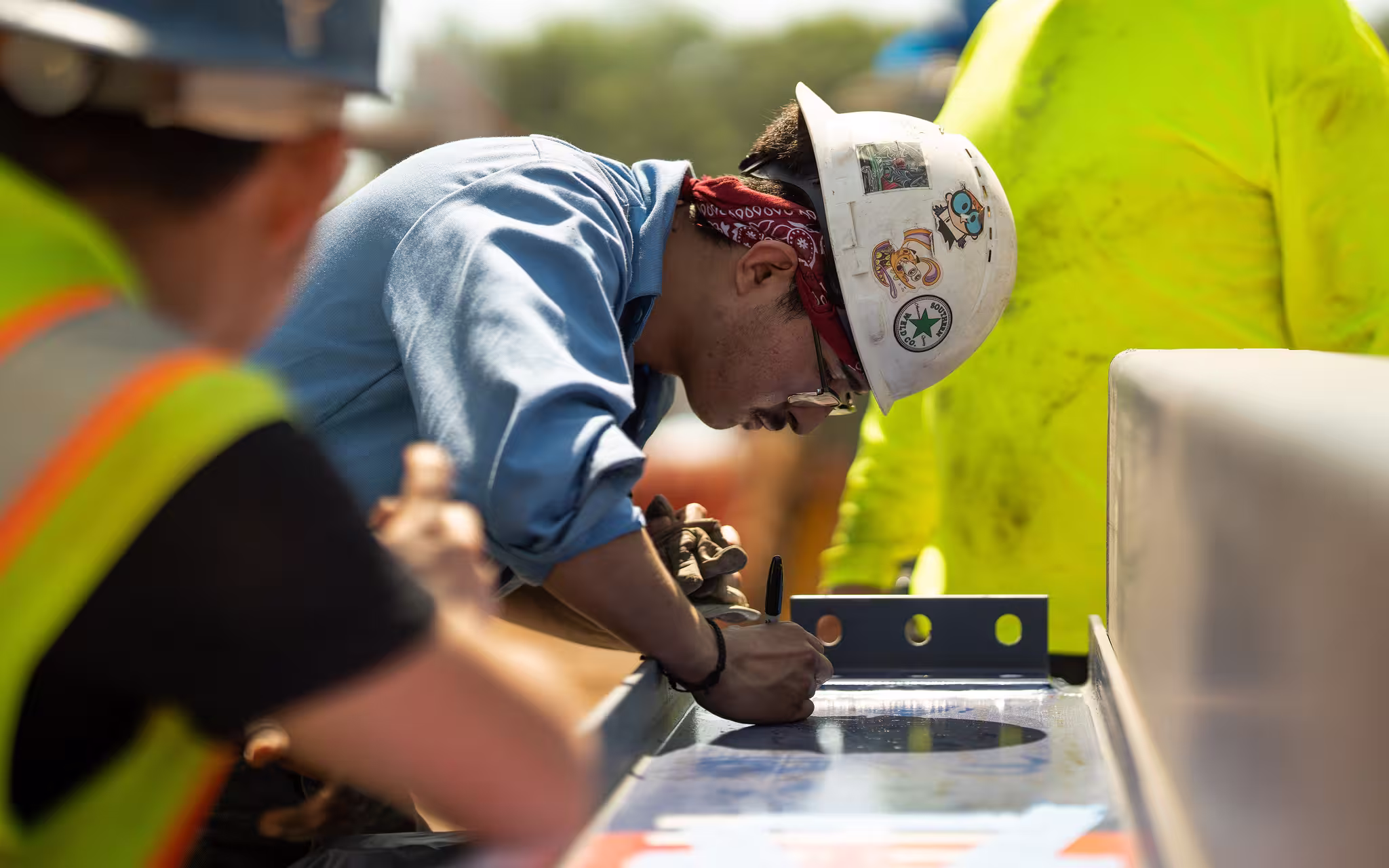 A construction worker wearing a sticker-covered hard hat marks a wooden beam, using a blue level as a guide.