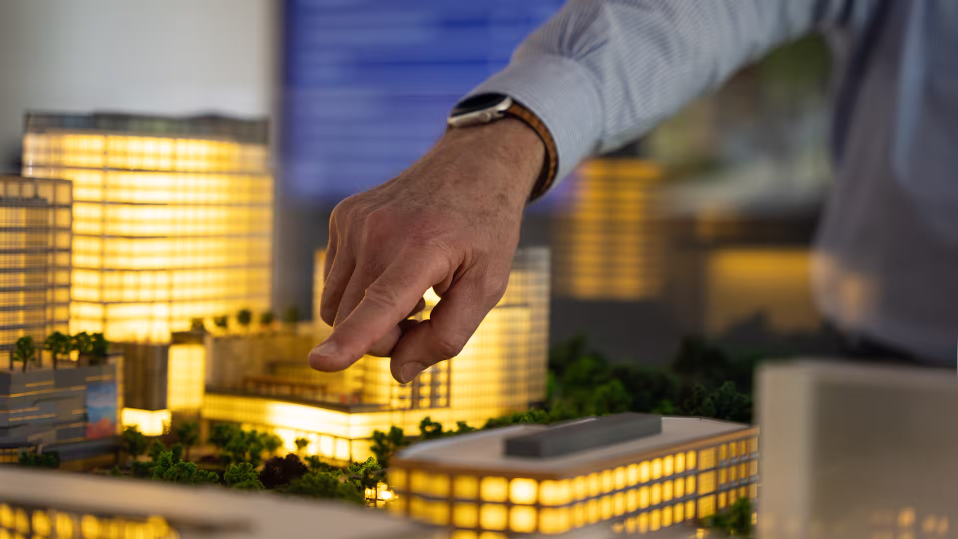 A close-up of a person's hand pointing at an illuminated architectural scale model of buildings.