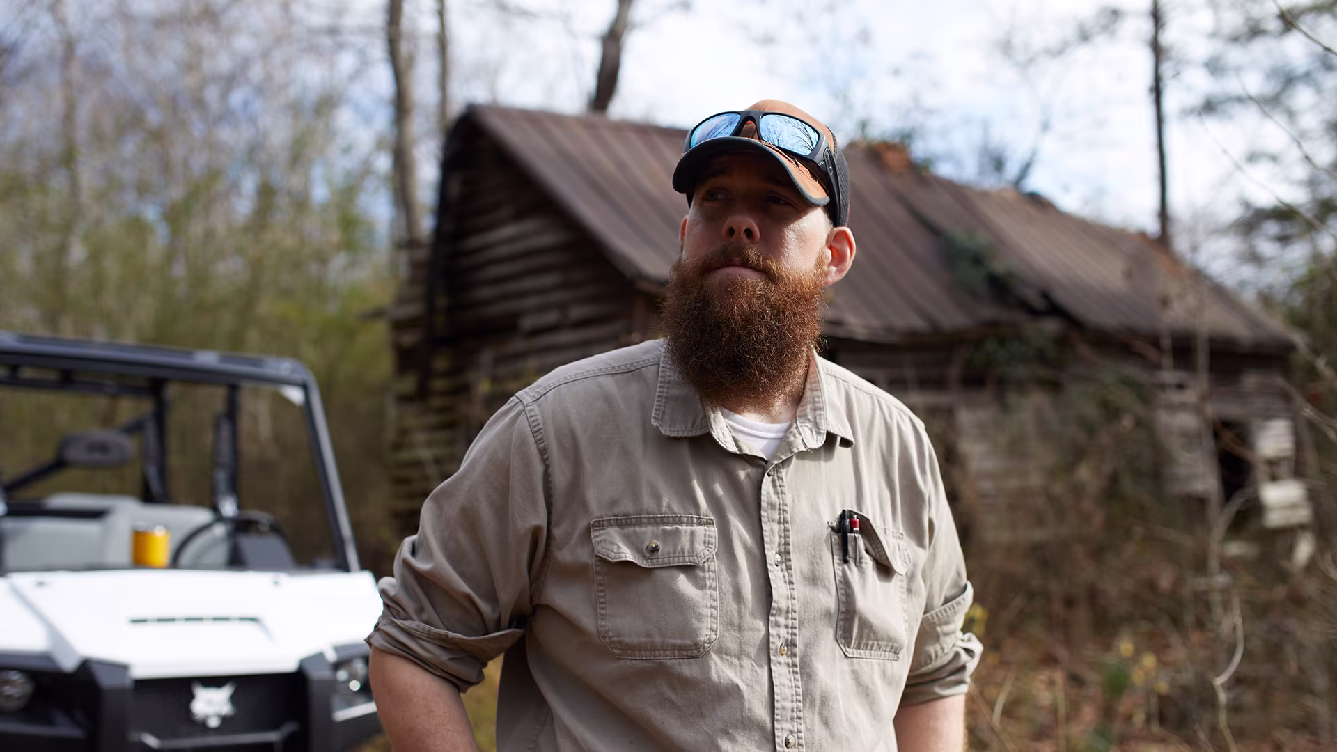 A bearded man wearing a khaki shirt and sunglasses on his head stands in a wooded area, looking upward. Behind him are a white utility vehicle and a dilapidated wooden cabin.