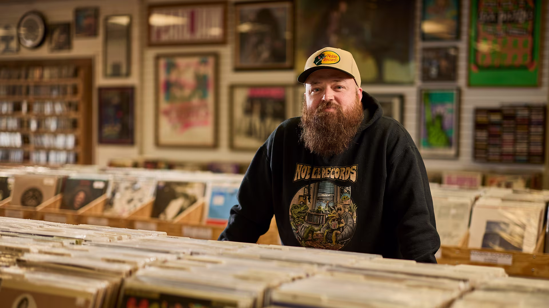 A bearded man in a cap and black hoodie stands behind rows of vinyl records inside a warmly lit record store.