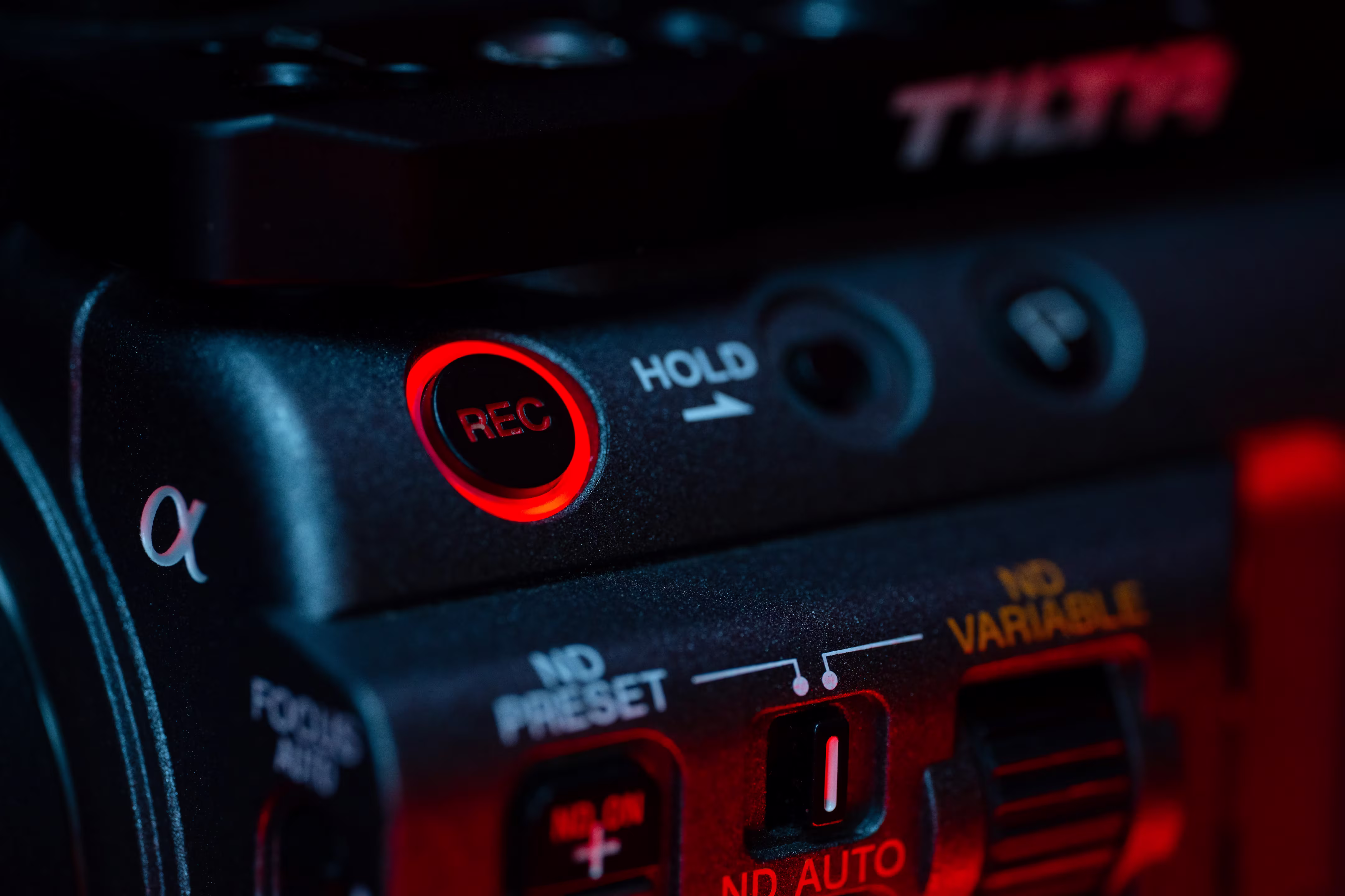 Close-up of a camera with a red glowing "REC" button, revealing brand markings and control dials, conveying a focused and technical atmosphere.