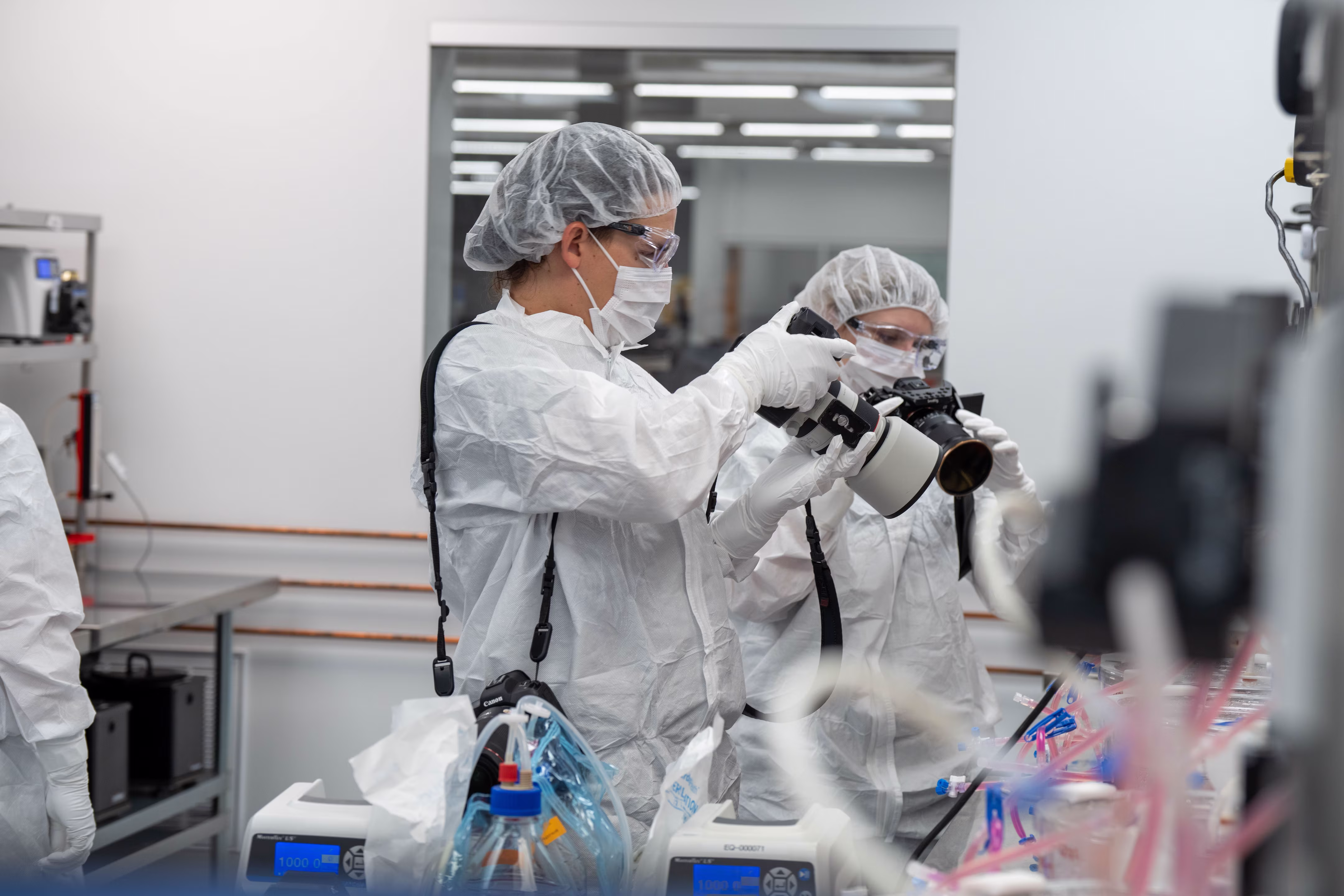 Two scientists in protective gear and masks work in a lab, handling equipment with care and concentration. The sterile environment is brightly lit.