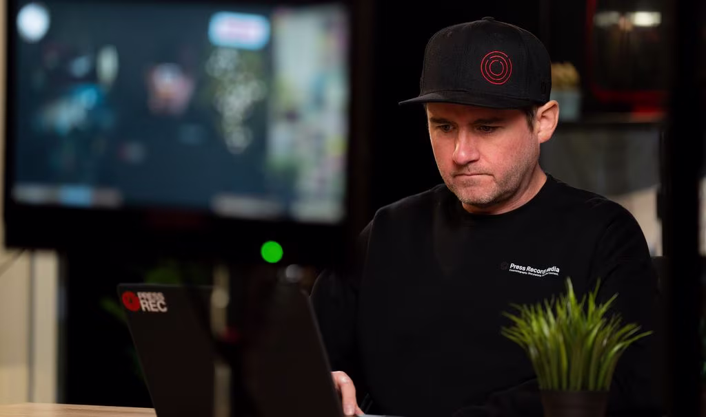 A man in a black cap works intently on a laptop at a desk. A monitor with blurred content is in the background. A small green plant is in the foreground.