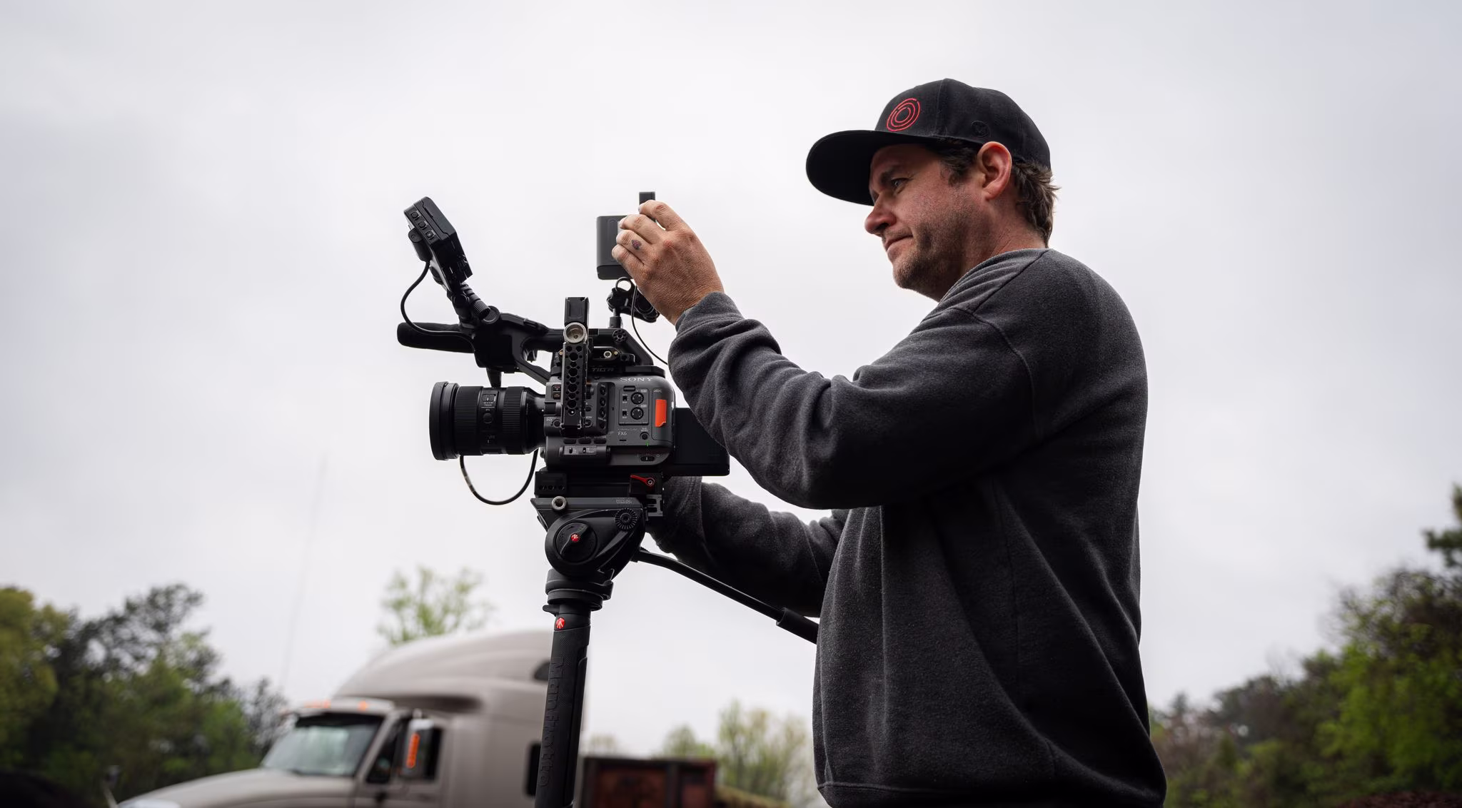 A man in a dark sweatshirt and cap operates a professional camera outdoors, focusing intently. Overcast sky and trees in the background suggest a calm setting.