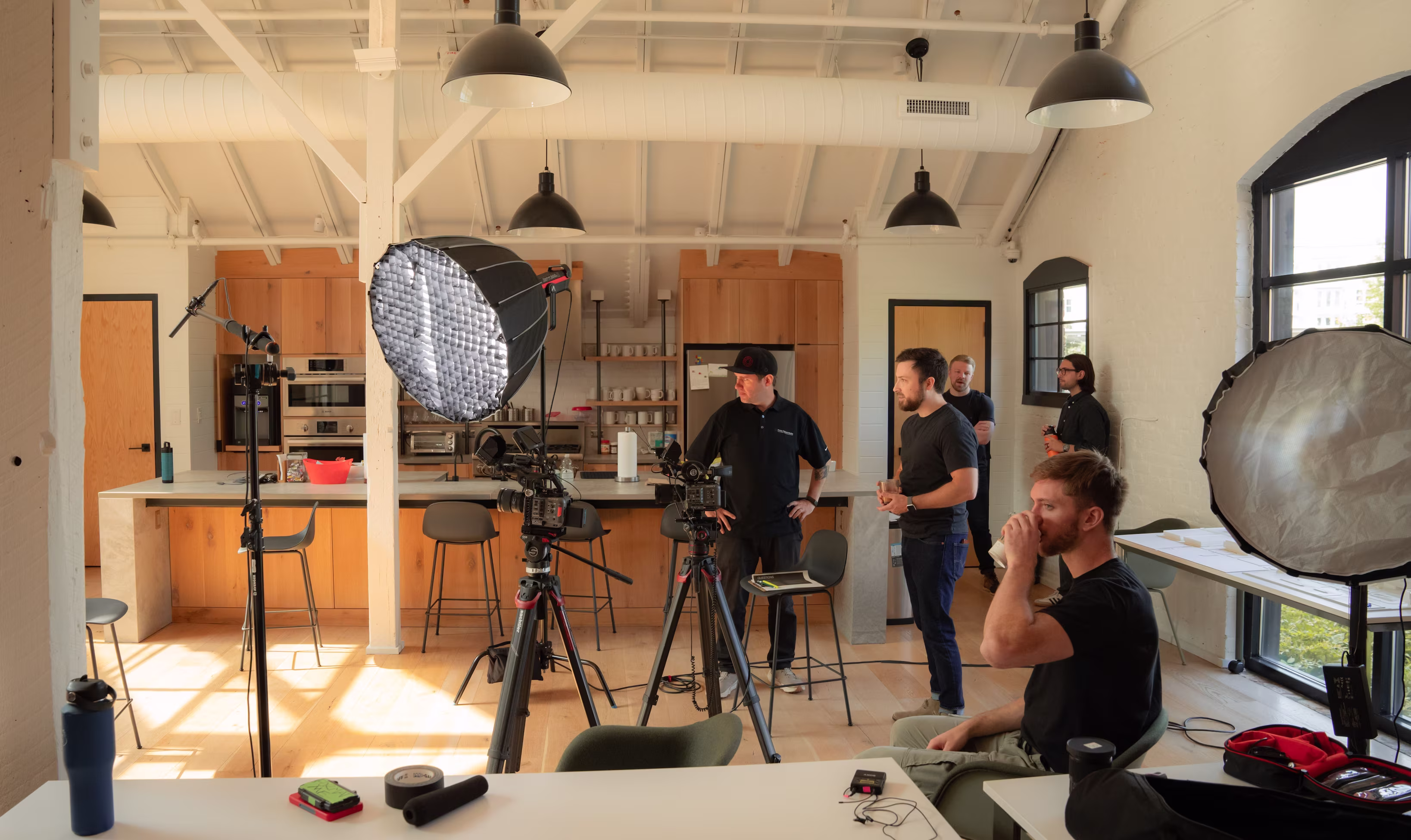 A busy film set in a modern kitchen with five crew members preparing equipment. Cameras, lights, and reflectors are arranged, creating a focused atmosphere.
