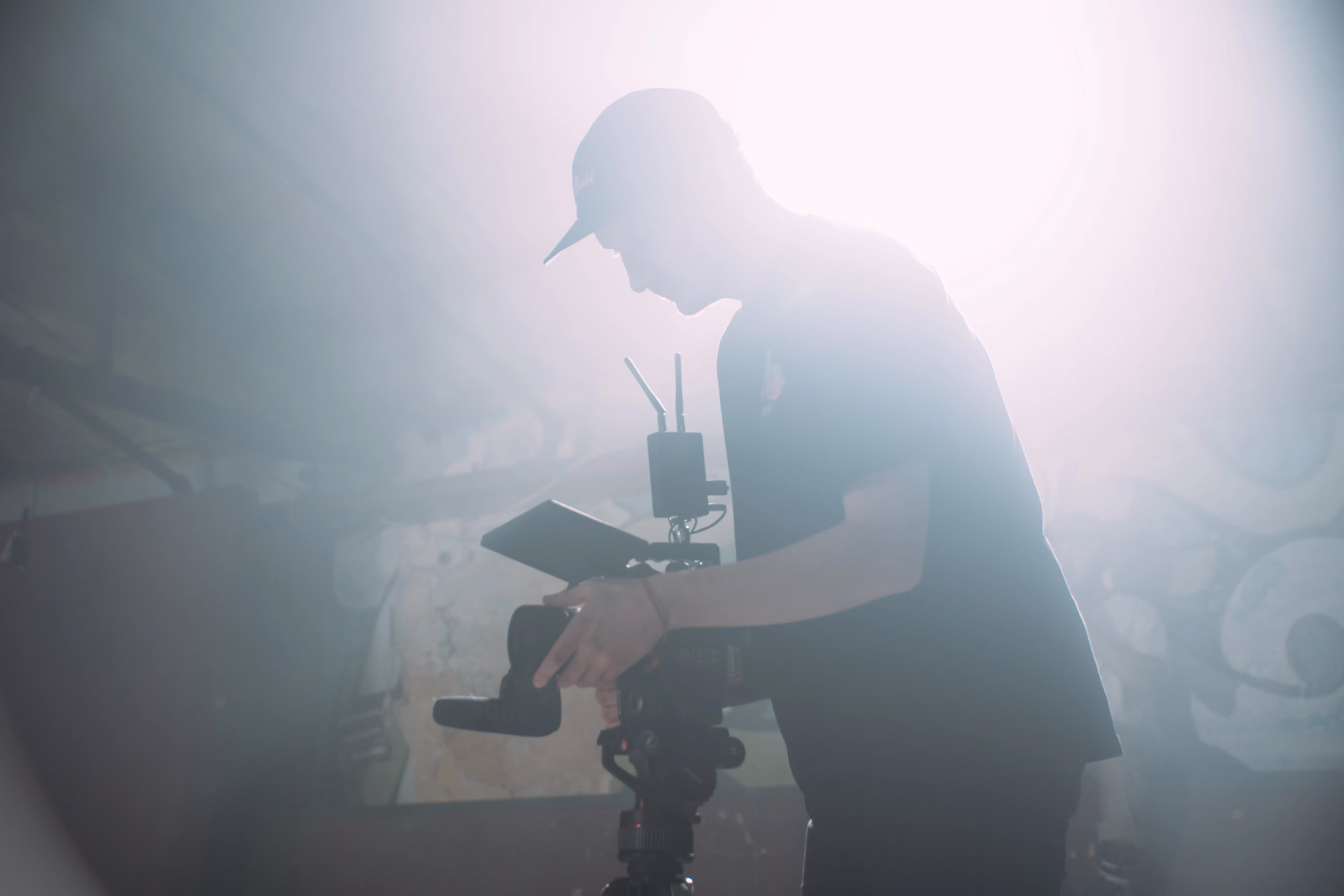 A silhouette of a person in a cap using a camera on a tripod in a dimly lit room. Bright backlighting creates a dramatic, atmospheric effect.