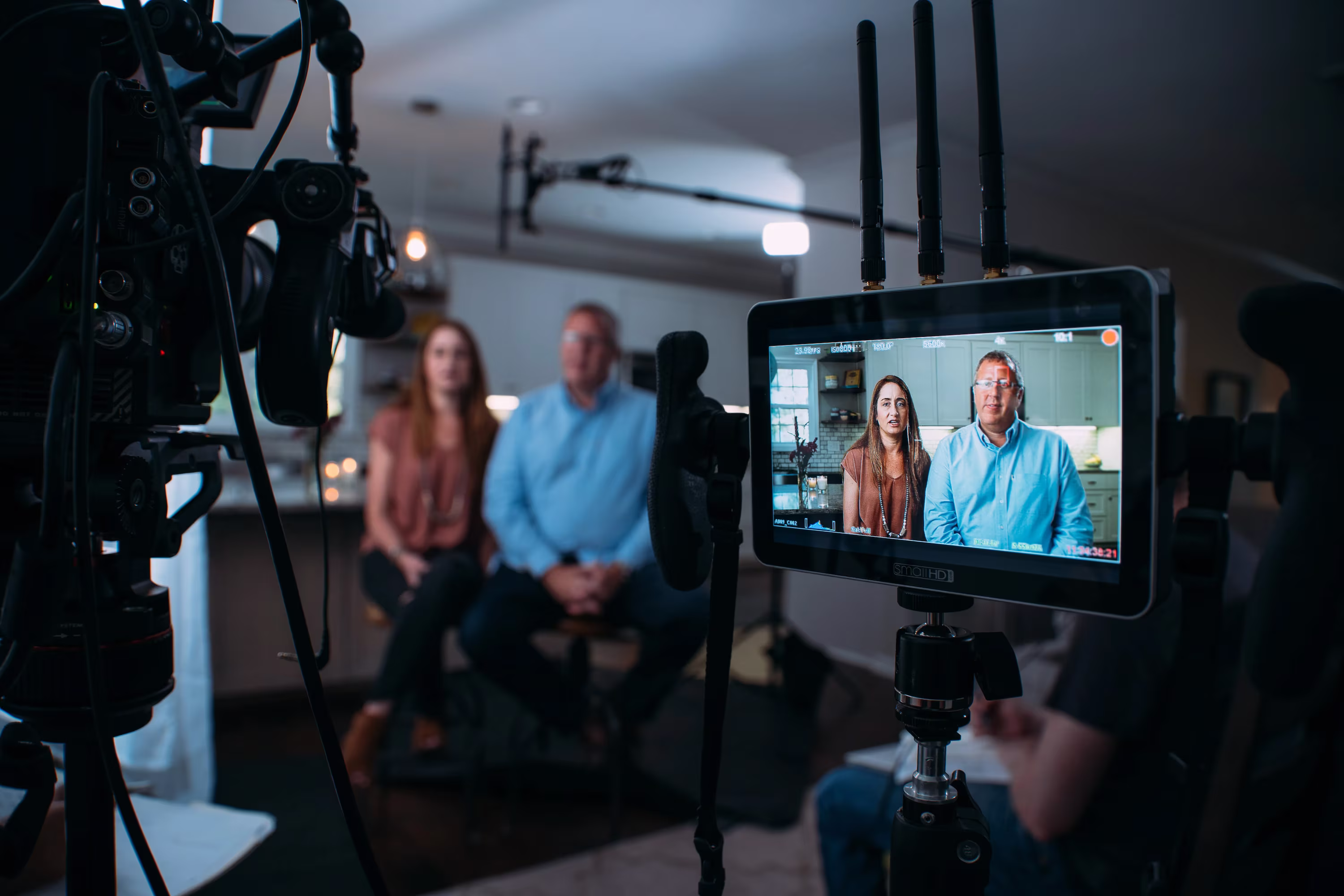 A woman and man sit together in a kitchen, framed in a camera monitor during a filmed interview setup. The atmosphere appears professional.