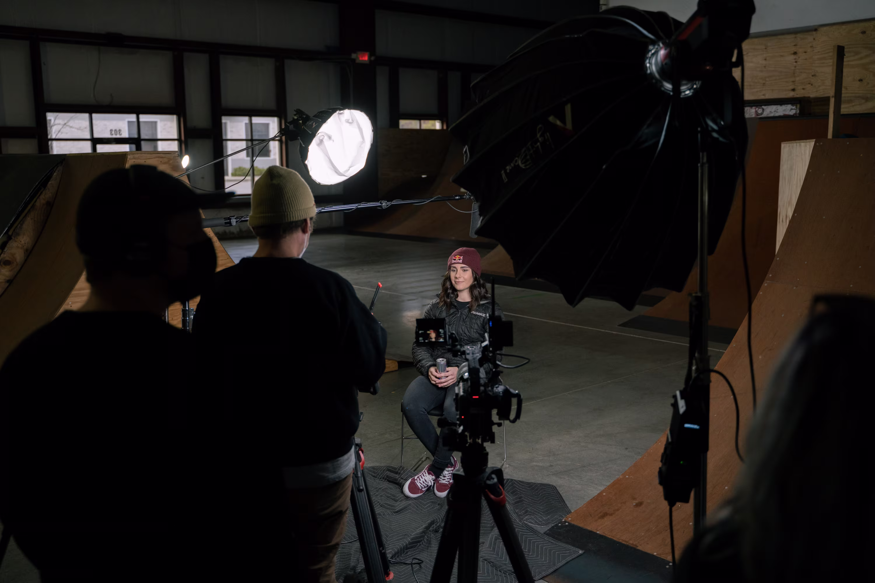 A person with a red beanie is being filmed in a dimly lit skatepark. Bright studio lights and cameras surround them, creating a focused interview setting.