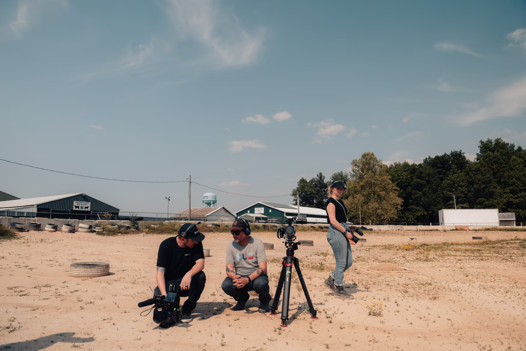 Three people with cameras on a sandy surface under a clear sky. Two crouch near a tripod; one stands, suggesting a filmmaking scene.