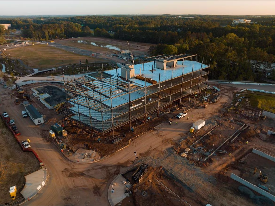 Aerial view of a large construction site with a steel-framed building in progress. Surrounding machinery and dirt roads create an industrious scene.