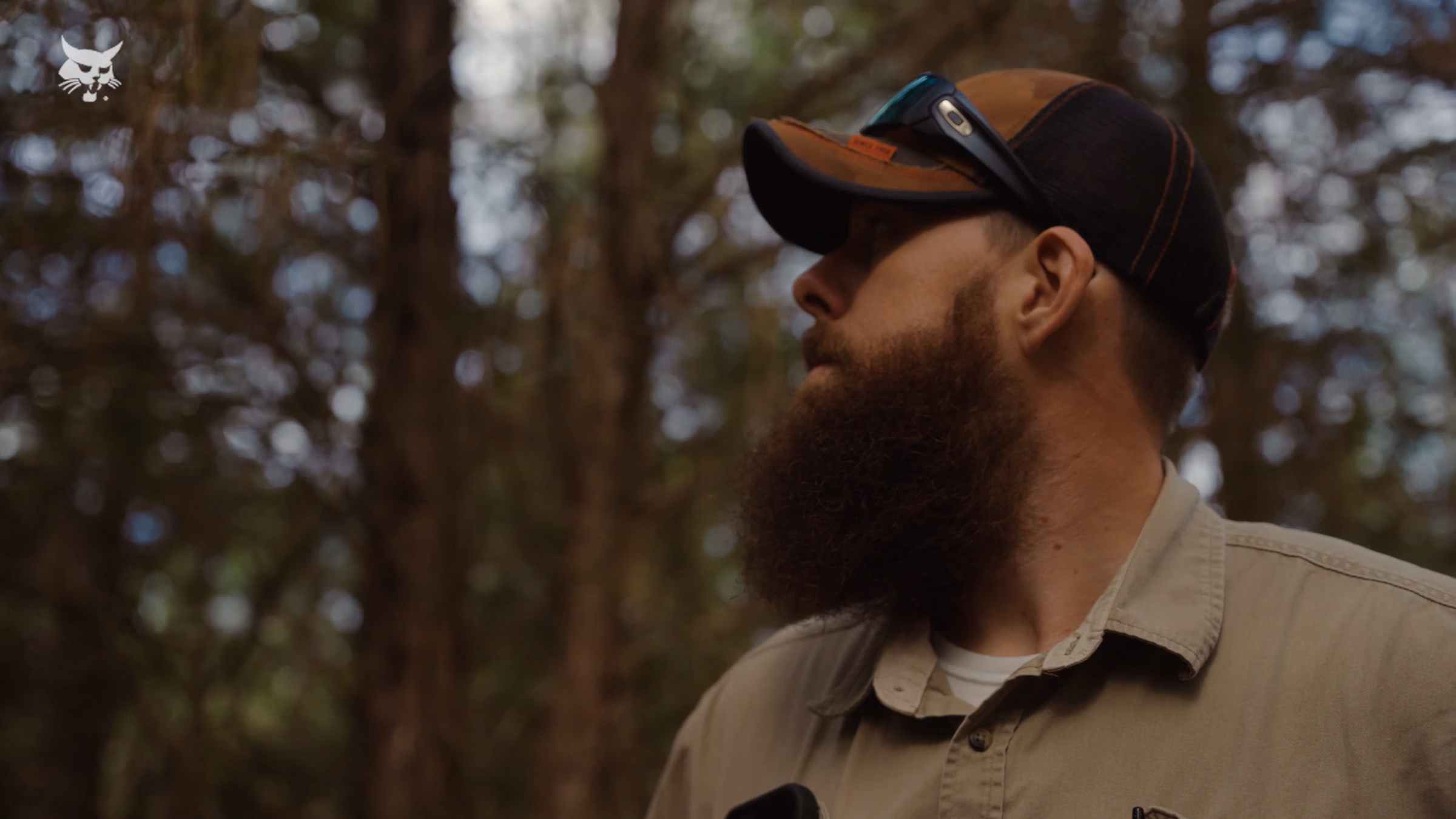 Bearded man wearing cap and sunglasses on head looks to the side in a wooded outdoor setting.