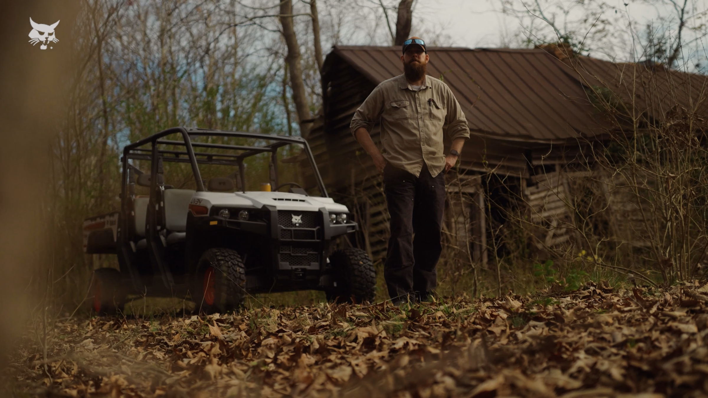 Bearded man in outdoor work clothes stands near utility vehicle and old wooden cabin in forest.